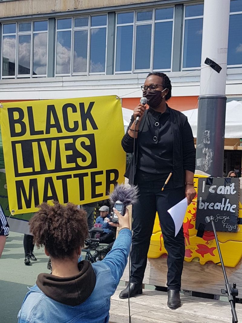 Das Bild zeigt Awet Tesfaiesus in schwarzer Kleidung mit einer Corona-Maske über dem Mund und einem Mikrofon in der Hand. Sie steht neben einem gelben Transparent mit der Aufschrift "BLACK LIVES MATTER". In der anderen Hand hält sie ein Blatt Papier. Tesfaiesus steht auf einer Bank und hält eine Rede. Vor ihr steht eine Person mit krausen Haaren, die Awet ein Mikrofon entgegenhält.