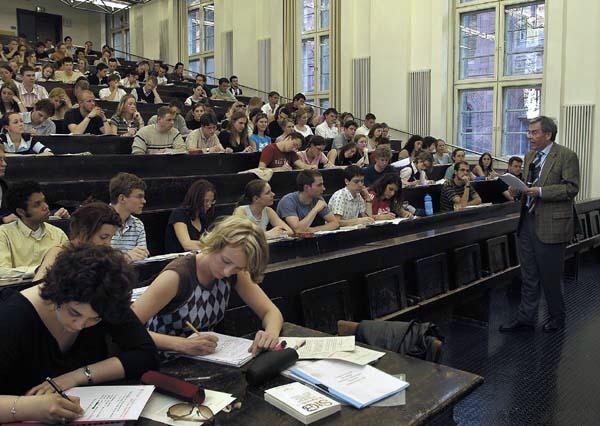 Das Bild zeigt den Hörsaal der juristischen Fakultät der Uni Heidelberg: auf steil aufsteigenden Bänken sitzen junge Menschen. Vorne steht ein älterer Mann im Anzug. Hält einen Stapel Blätter in den hängen. Es ist der Professor. Im Vordergrund liegt ein Buch vor zwei Studierenden auf einem Tisch. Darauf steht: StGB.