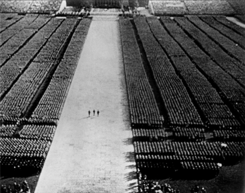 Das Bild zeigt eine Szene aus Leni Riefenstahls Film "Der Triumph des Willens". Zu sehen ist das Gelände des Reichsparteitages aus der Vogelperspektive. Drei Figuren schreiten eine breite Mittelbahn ab. Rechts und links von Ihnen stehen gewaltige Kolonnen von Soldaten in Reih und Glied.